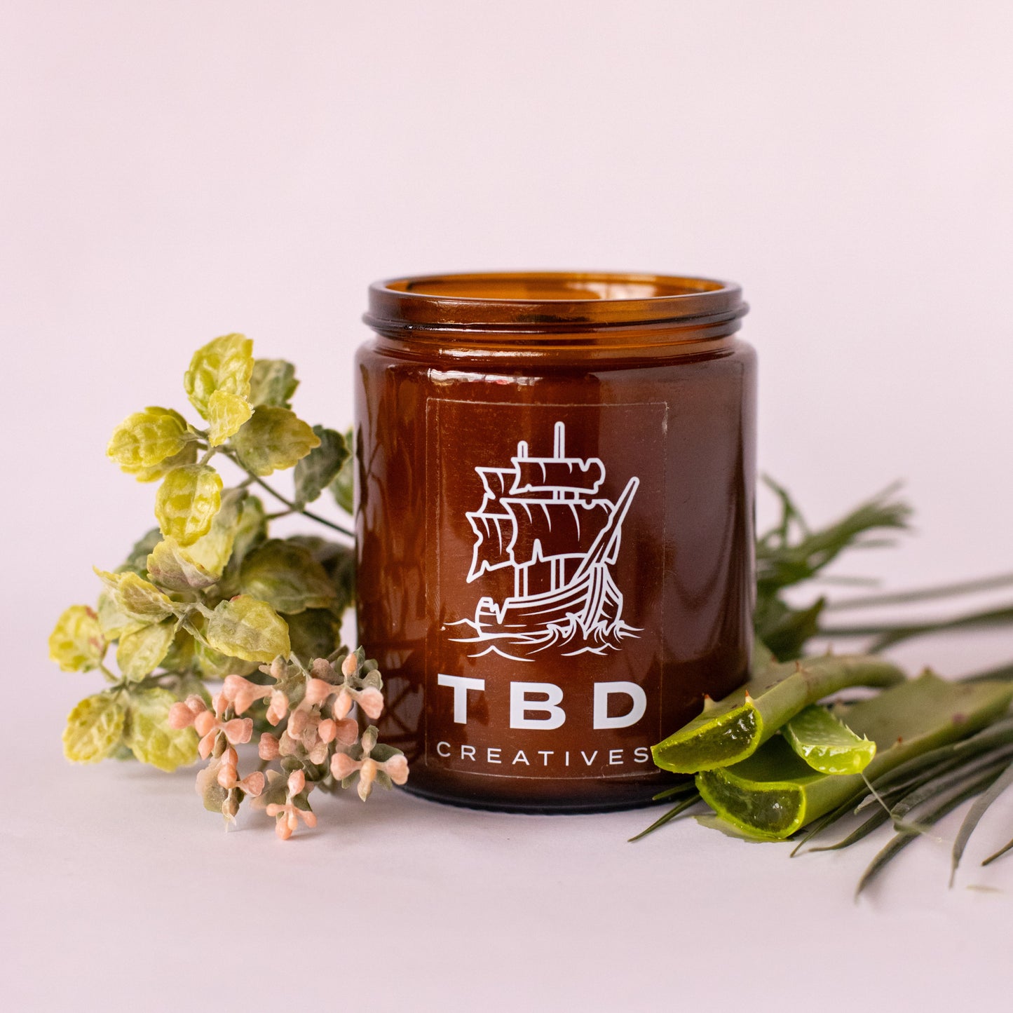 Brown jar with 'TBD Creatives' logo on a light pink background with greenery.