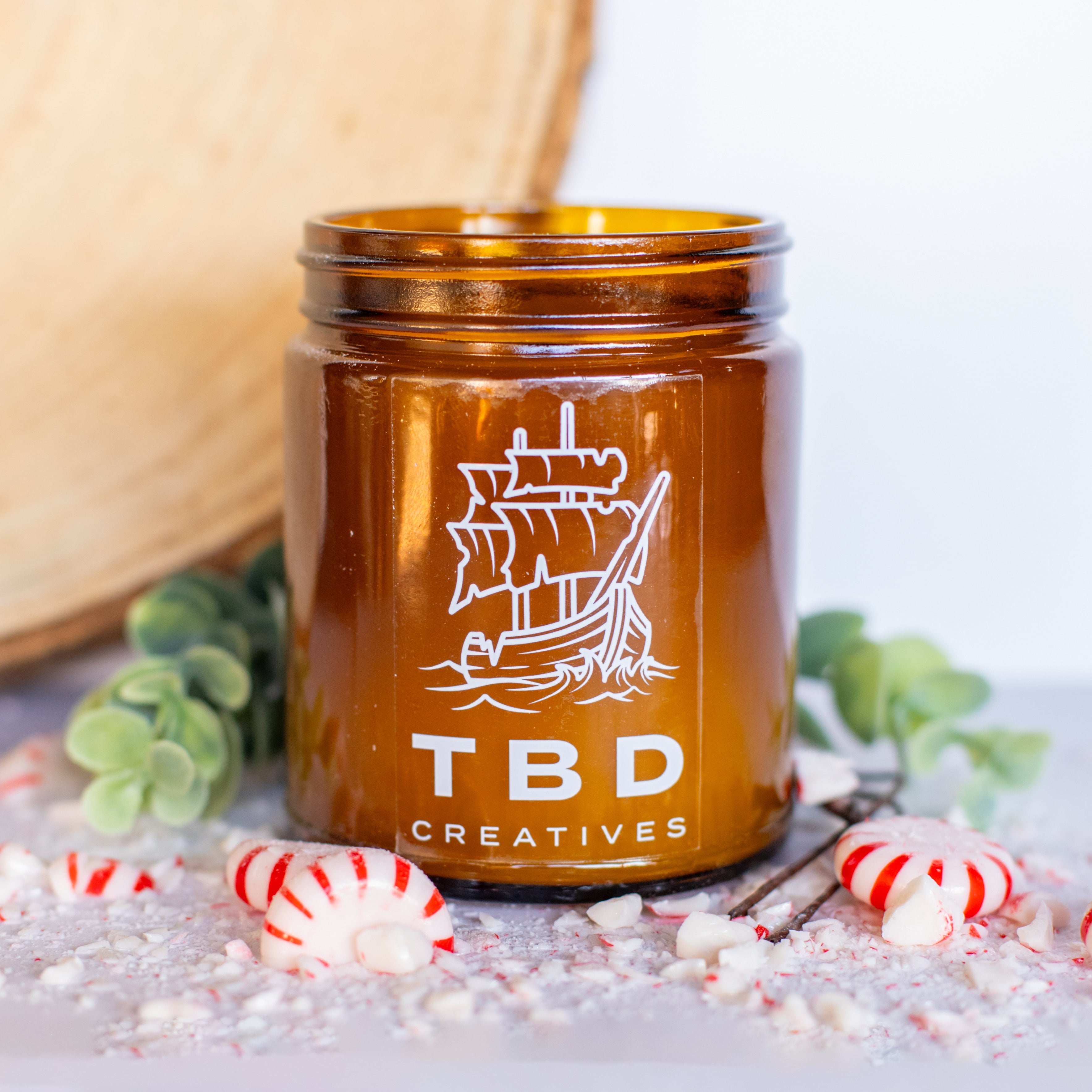Brown candle jar with ship design and 'TBD Creatives' text on a light background with candy and decorative elements.