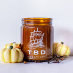 Amber glass jar with ship design and 'TBD Creatives' text, surrounded by small pumpkins and cinnamon sticks on a light background.