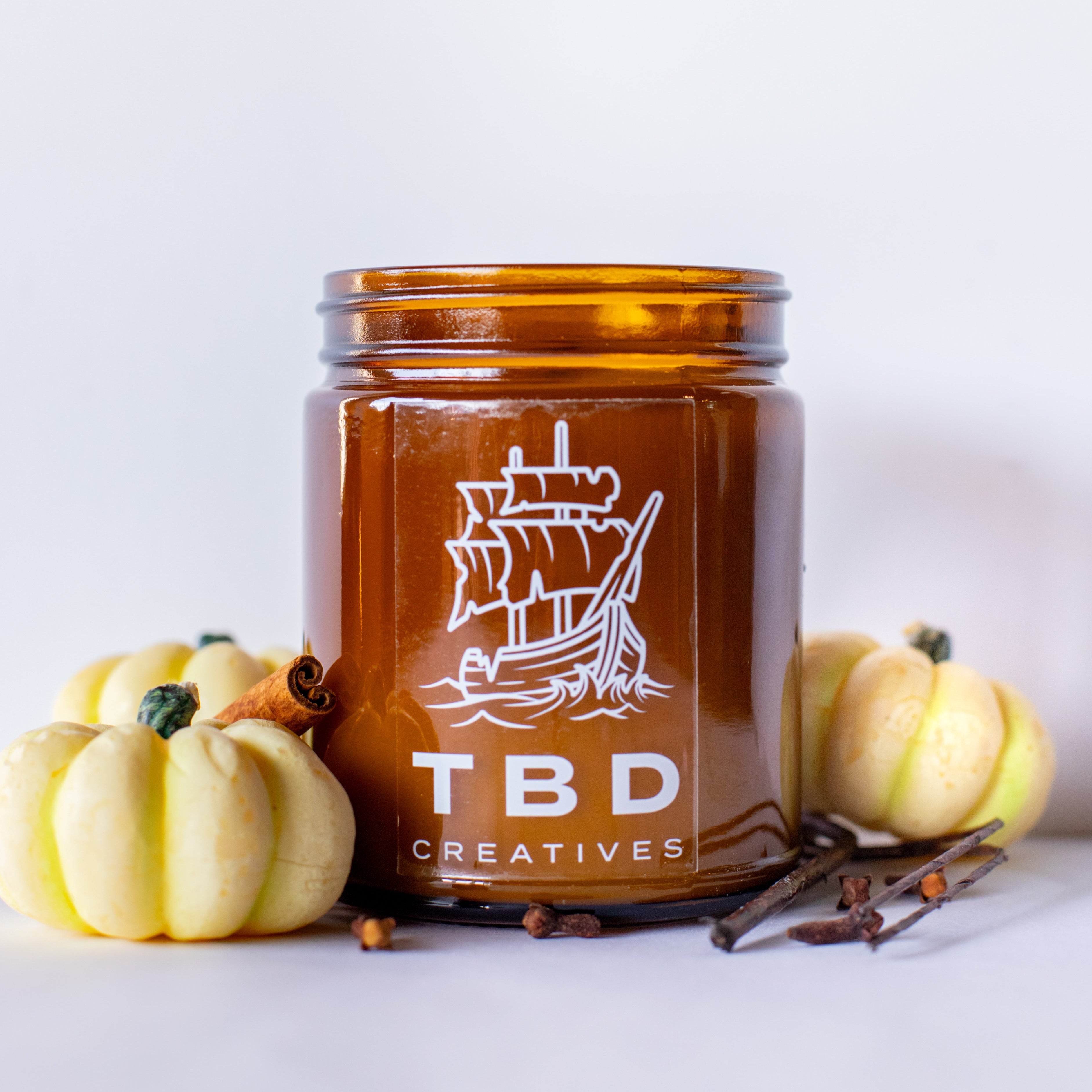 Amber glass jar with ship design and 'TBD Creatives' text, surrounded by small pumpkins and cinnamon sticks on a light background.