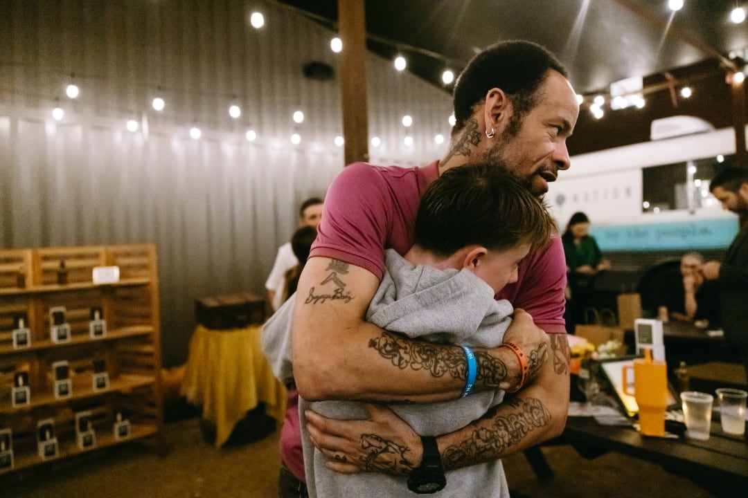 Two people hugging in a casual indoor setting with shelves and people in the background.
