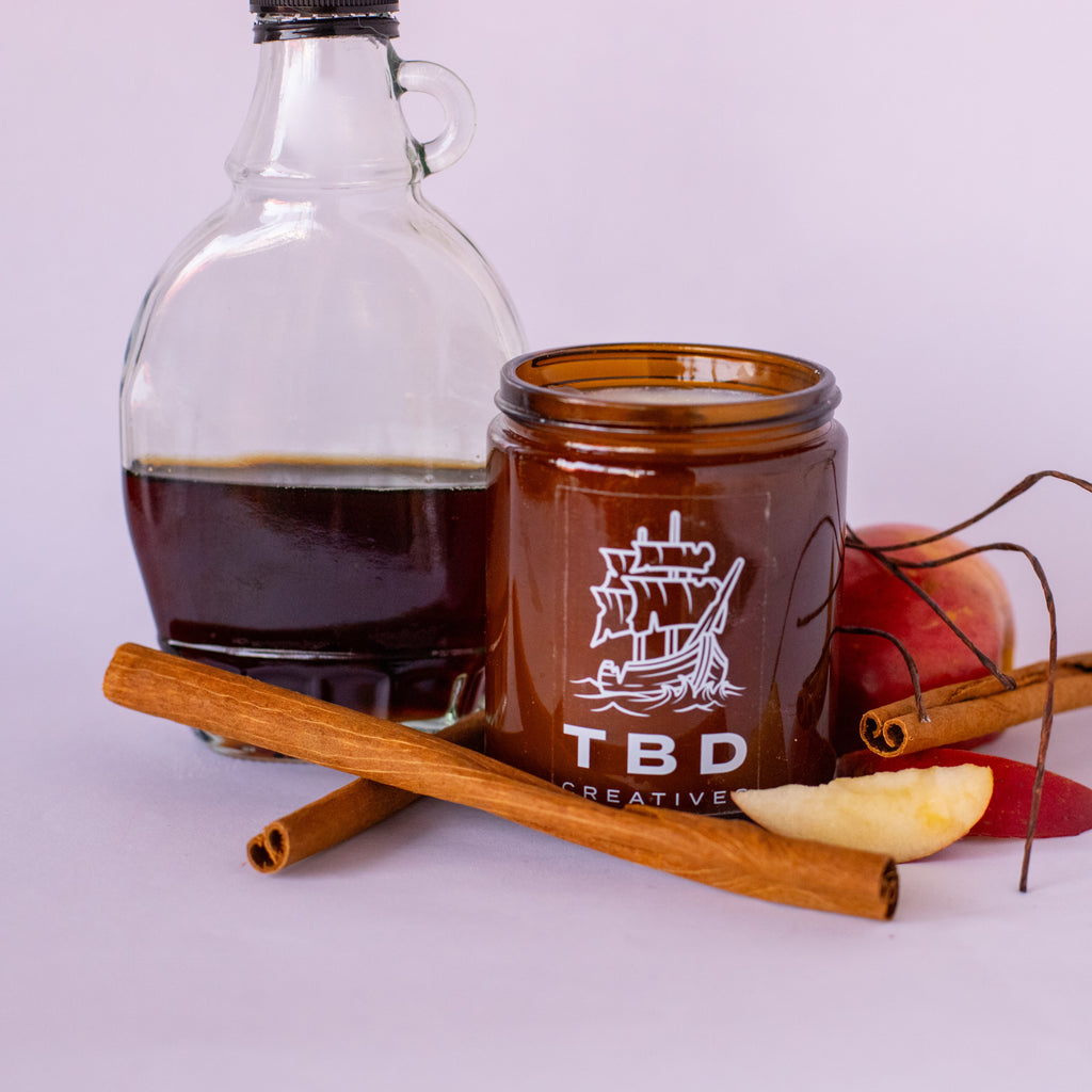 Jar with 'TBD Creative' branding surrounded by cinnamon sticks, apples, and a bottle on a light background