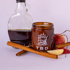 Jar with 'TBD Creative' branding surrounded by cinnamon sticks, apples, and a bottle on a light background