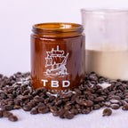 Amber glass jar with ship logo and 'TBD' text on a white background with coffee beans.