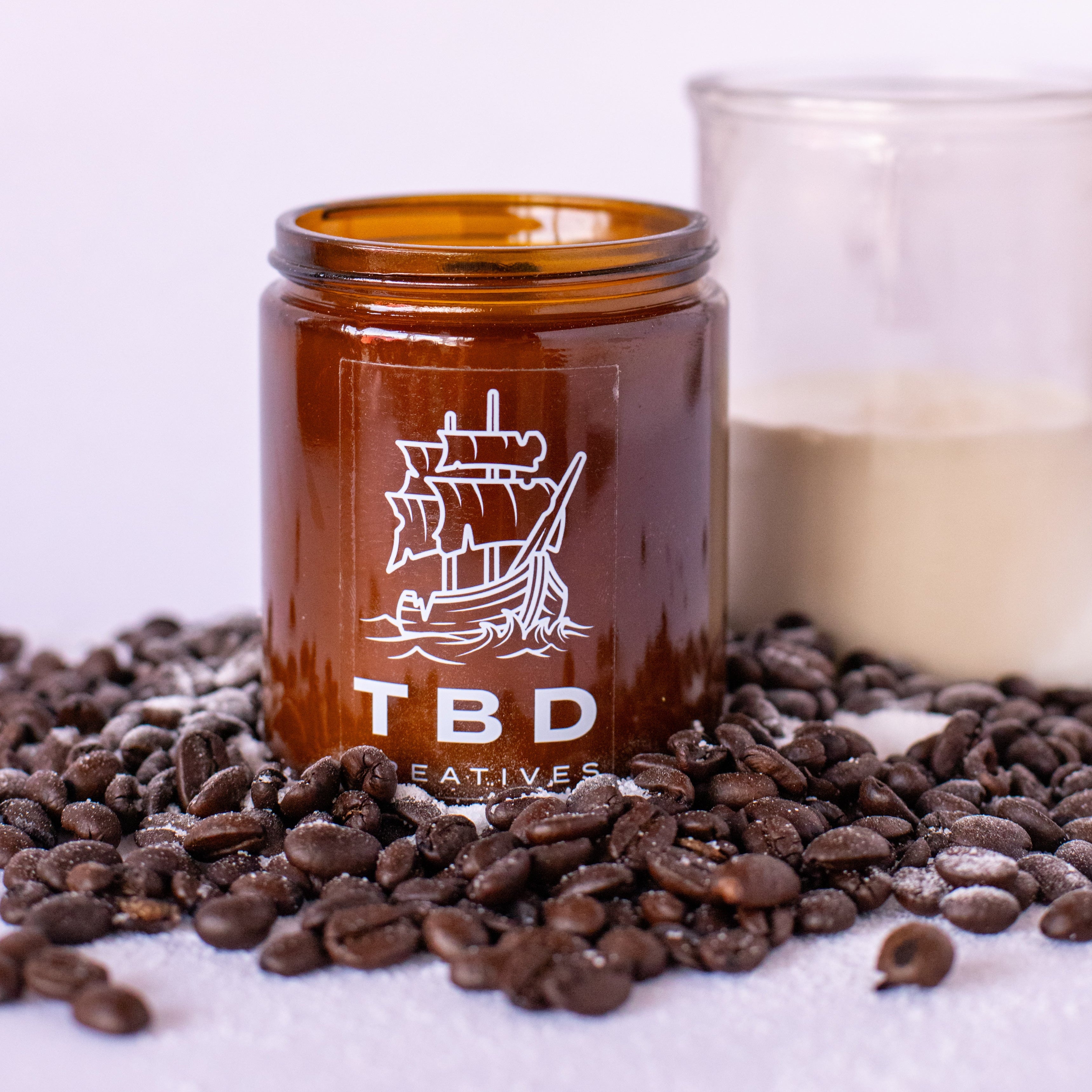 Amber glass jar with ship logo and 'TBD' text on a white background with coffee beans.
