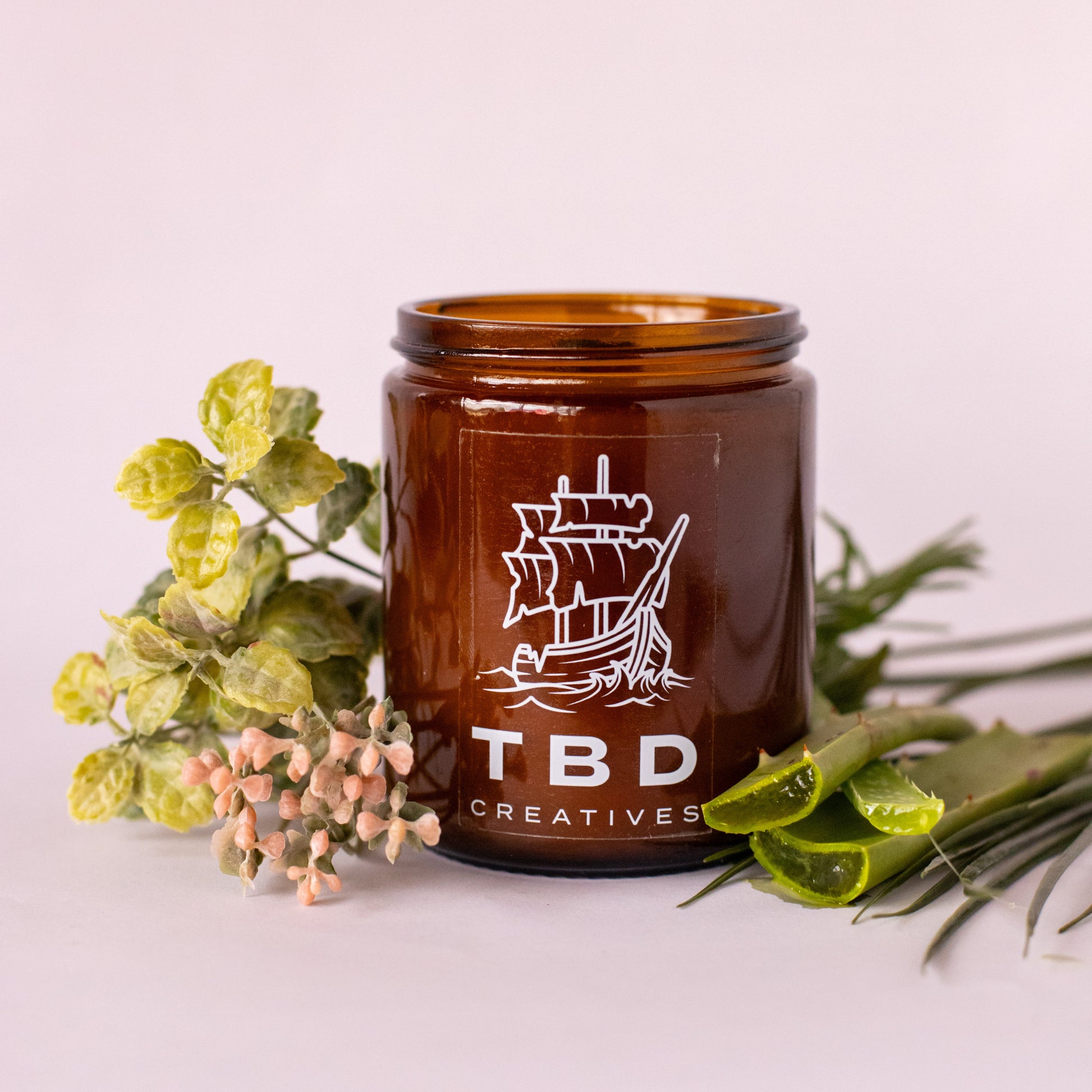 Brown jar with 'TBD Creatives' logo on a light pink background with greenery.