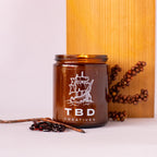 Brown jar with 'TBD Creatives' logo on a white background with wooden board and coffee beans.