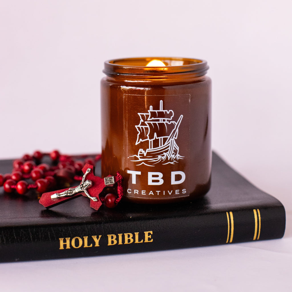Brown candle with 'TBD Creatives' logo on a black book titled 'HOLY BIBLE'.