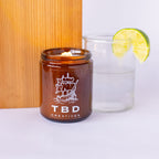 Amber candle in a glass jar with 'TBD Creatives' branding, next to a glass of water and lime wedge on a white surface.