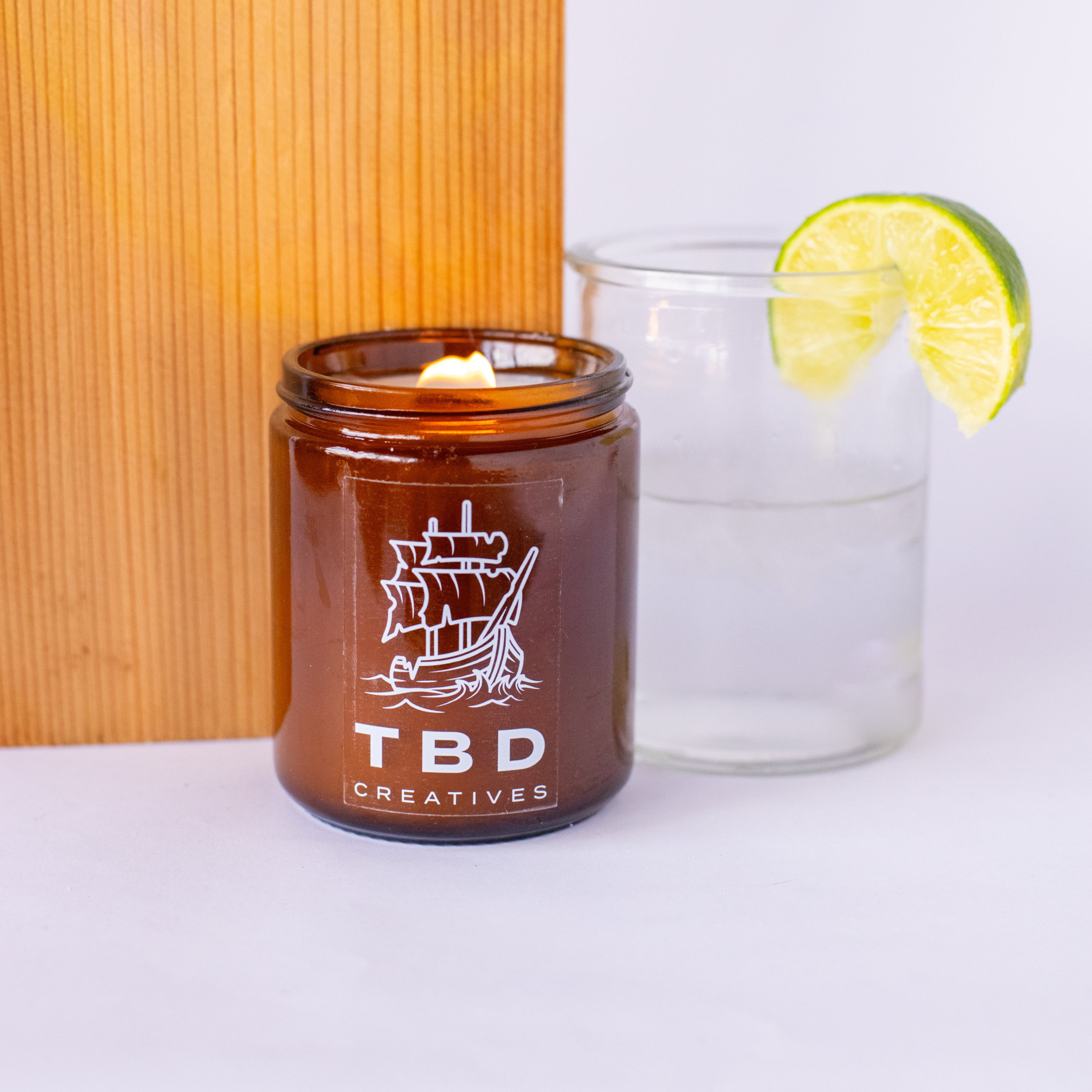 Amber candle in a glass jar with 'TBD Creatives' branding, next to a glass of water and lime wedge on a white surface.