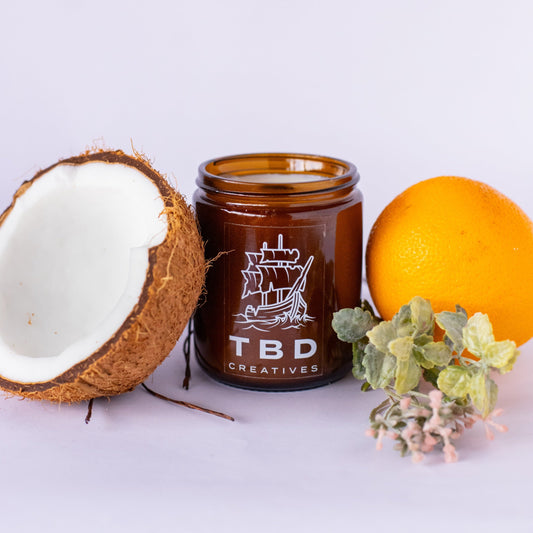 Brown jar with 'TBD Creatives' branding, half-coconut, orange, and small plant on a light background