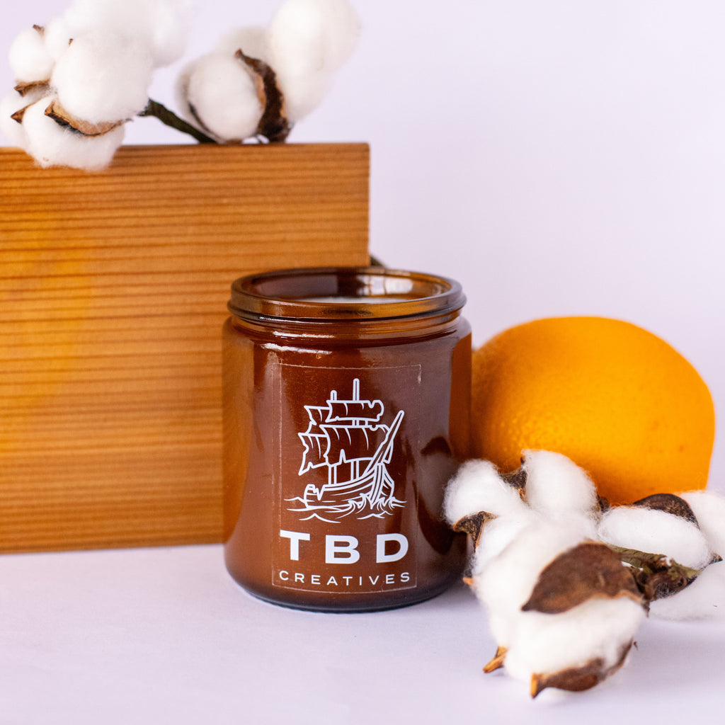 Brown candle with 'TBD Creatives' logo on a white surface with cotton and wooden block in the background
