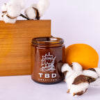 Brown candle with 'TBD Creatives' logo on a white surface with cotton and wooden block in the background