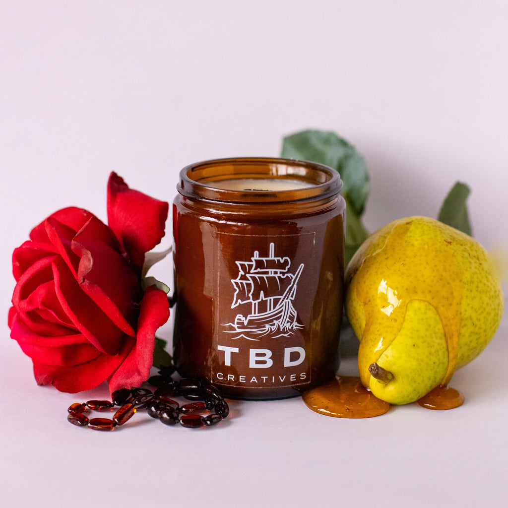Jar of candle with 'TBD Creatives' branding, surrounded by a red rose, green leaf, and yellow fruit on a light background.