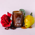 Jar of candle with 'TBD Creatives' branding, surrounded by a red rose, green leaf, and yellow fruit on a light background.