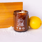 Brown candle jar with 'TBD Creatives' logo next to a lemon and decorative box on a white background