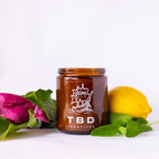 Jar with 'TBD Creatives' branding on a white background with pink flower and lemon