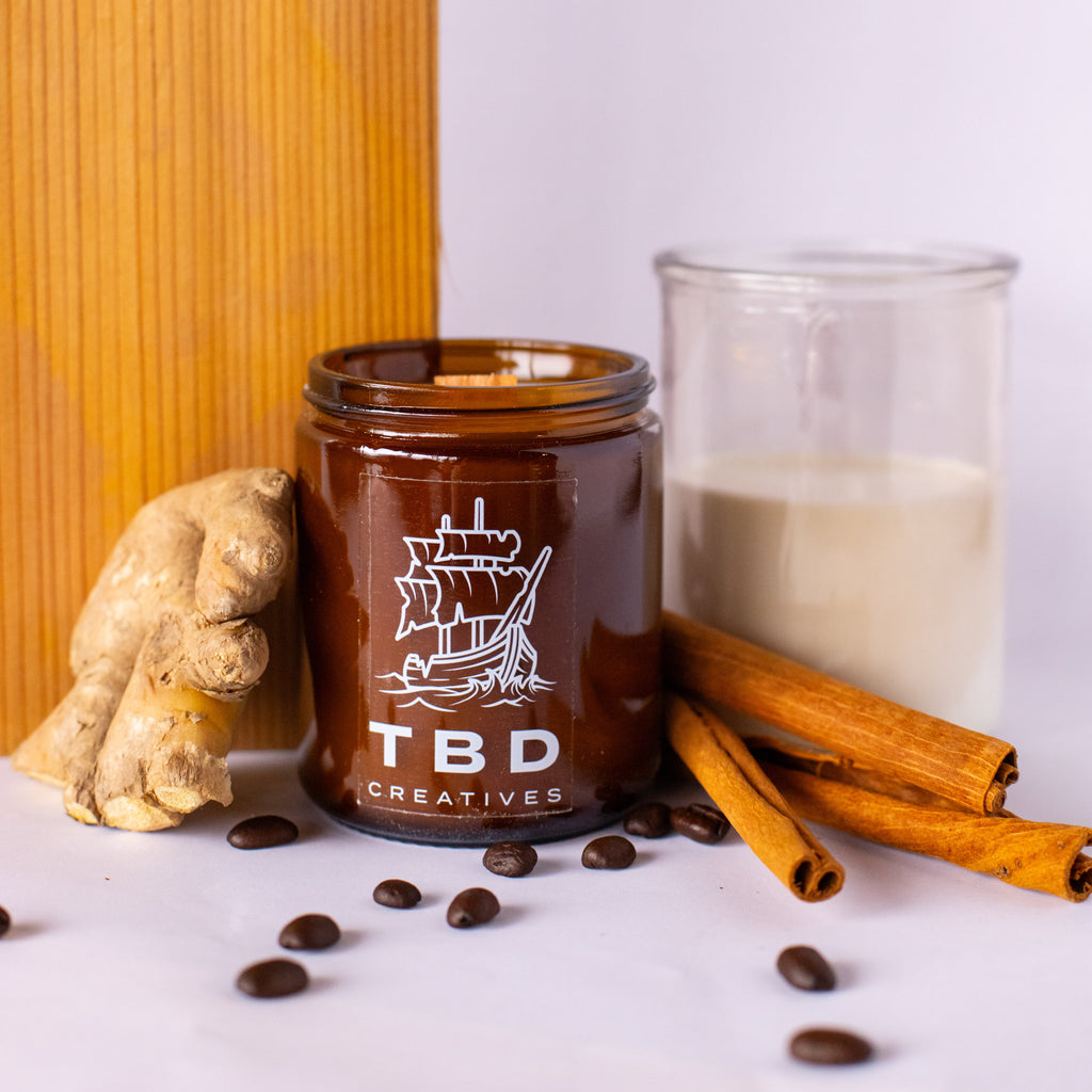 Brown candle jar with 'TBD Creatives' branding, ginger root, cinnamon sticks, and coffee beans on a white surface.